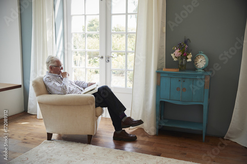 Senior man sitting in an armchair reading a book at home © Monkey Business