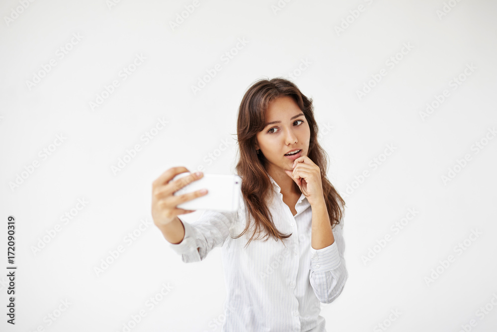 Collecting memories. Headshot of charming young woman employee in formal wear taking self pictures using smart phone. Beautiful Latin female worker posing at white office wall, taking selfie Collecting memories. Headshot of charming young woman employee in formal wear taking self pictures using smart phone. Beautiful Latin female worker posing at white office wall, taking selfie