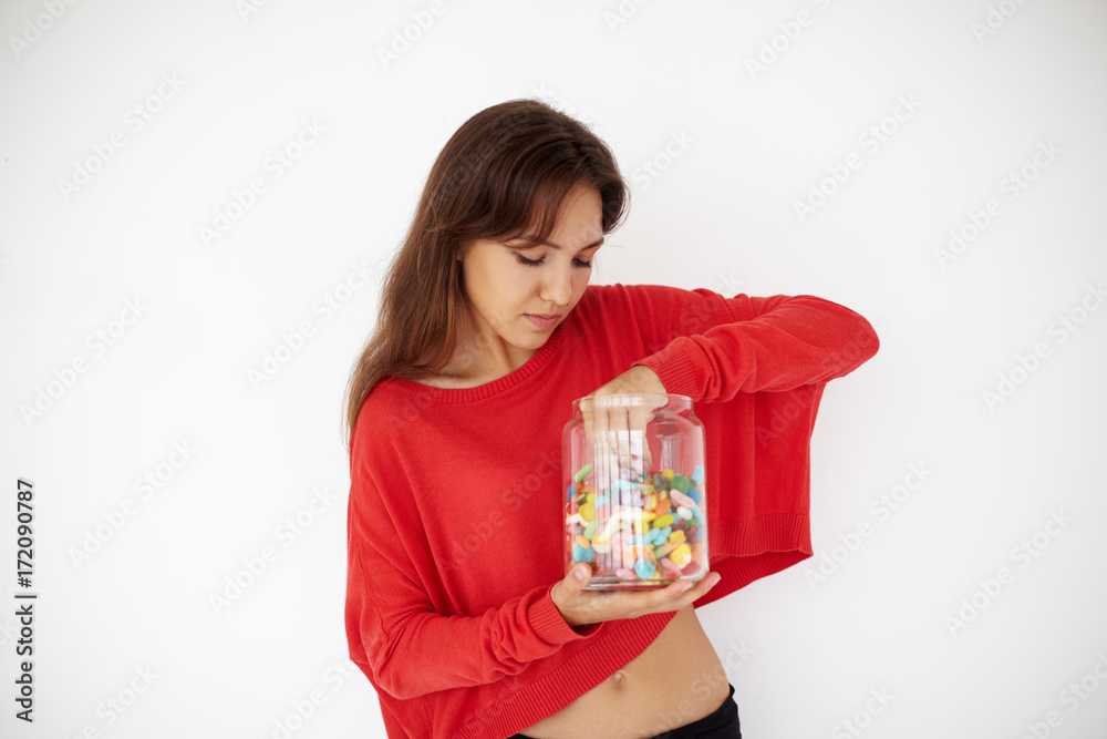 Horizontal studio shot of casually dressed young brunette woman picking out dragee, holding glass jar, having impatient expression. Cute teenage girl in red sweater eating colorful sweets after lunch Horizontal studio shot of casually dressed young brunette woman picking out dragee, holding glass jar, having impatient expression. Cute teenage girl in red sweater eating colorful sweets after lunch