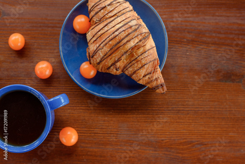 Morning breakfast: blue cup of tea, chocolate croissant and physalis on a wooden table