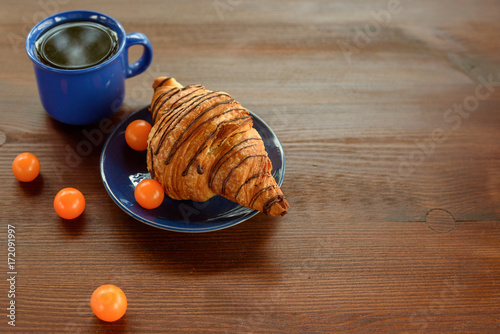 Morning breakfast: blue cup of tea, chocolate croissant and physalis on a wooden table