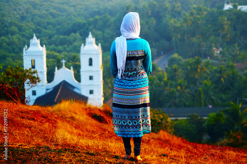 Young christian female in front of the church on the hill. Scarf on her head. She's in traditional indian clothes.