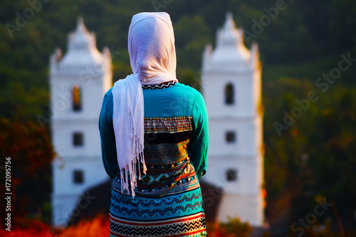 Young christian female in front of the church on the hill. Scarf on her head. She's in traditional indian clothes.