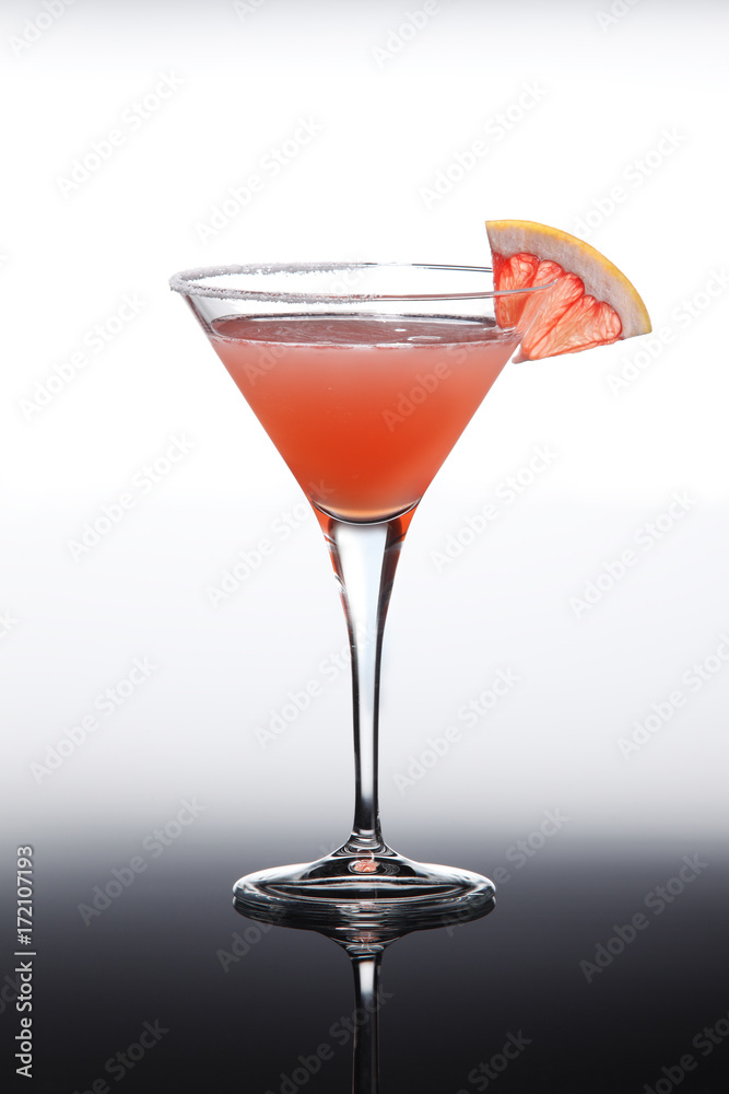 Cocktail with cranberry and grapefruit juice topped with grapefruit slice, in martini glass. On gradient background with reflection.