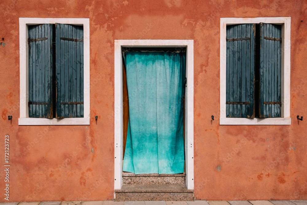 Beautiful colorful house facade on Burano island, north Italy. Red old house wall with a door covered with azure cloth and windows