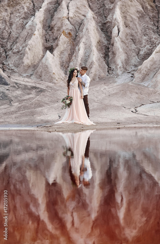 Fototapeta Naklejka Na Ścianę i Meble -  Couple in love hugging against the background of mountains and red lake. Unrealistic fairy-tale landscape, the mountains of Mars. Woman with a bouquet of flowers in the arms of men