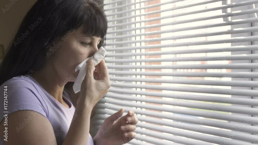 A young beautiful woman stands near a window and wipes his nose. A cold girl has an attack of runny nose at home. Allergies, colds, flu, acute respiratory infections, infection. A young beautiful woman stands near a window and wipes his nose. A cold girl has an attack of runny nose at home. Allergies, colds, flu, acute respiratory infections, infection.