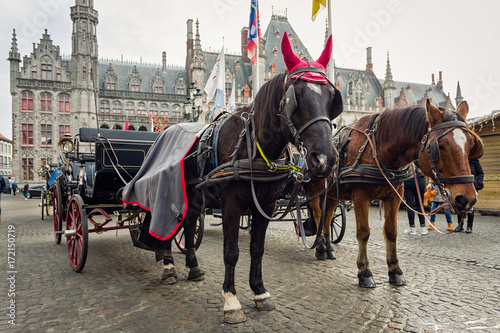 Horse-drawn carriage waiting on tourists for a guided tour on the center square in Bruges, Belgium. Christmas theme hat and ear covers on the horse, Christmas market on the background © danylamote