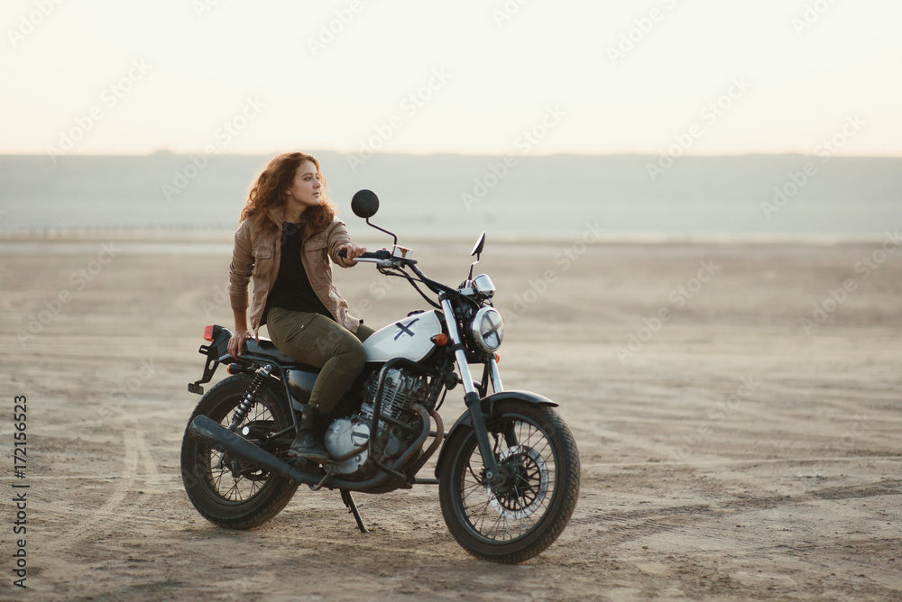 Obraz premium young beautiful woman sitting on her old cafe racer motorcycle in desert at sunset or sunrise
