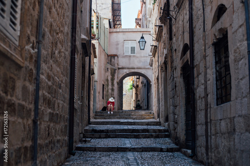 Fototapeta Naklejka Na Ścianę i Meble -  Narrow steep street of beautiful ancient european village or city, Dubrovnik Croatia with cobbled paved walls and floors. Lonely lost tourist sits on steps, exploring