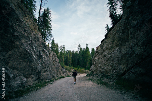 Curious explorer male figure of adventurer nomad traveler walks on path inbetween mountains and forest high in italian dolomites on sunset in evening