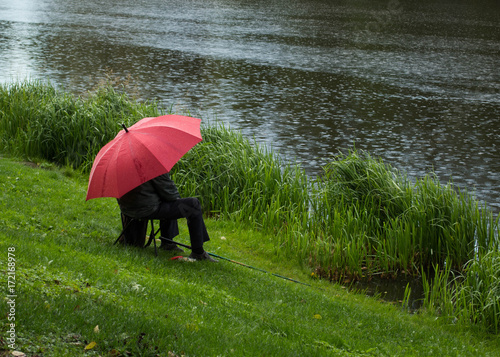  Senior man is sitting under a red umbrella and fishing on the lake bank on a autumn rainy day.