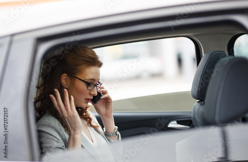 The red-haired female businesswoman, a distinguished boss, sits in the backseat of her car, engaged in a focused phone conversation, managing business matters with finesse. © BalanceFormCreative