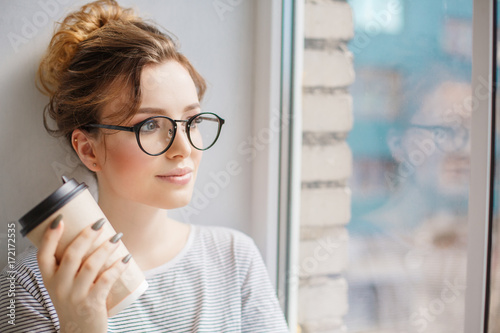 Young trendy woman by the window. A hipster style girl with a phone and coffee in fashion glasses sits alone on a windowsill