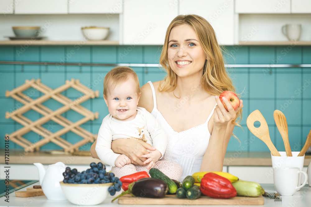 Young mother looking at camera and smiling, cooking and playing with her baby daughter in a modern kitchen setting. Healthy food concept.