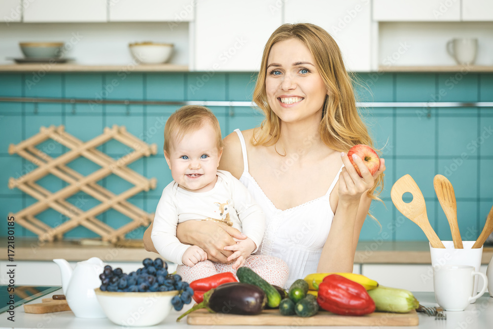 Young mother looking at camera and smiling, cooking and playing with her baby daughter in a modern kitchen setting. Healthy food concept. Young mother looking at camera and smiling, cooking and playing with her baby daughter in a modern kitchen setting. Healthy food concept.