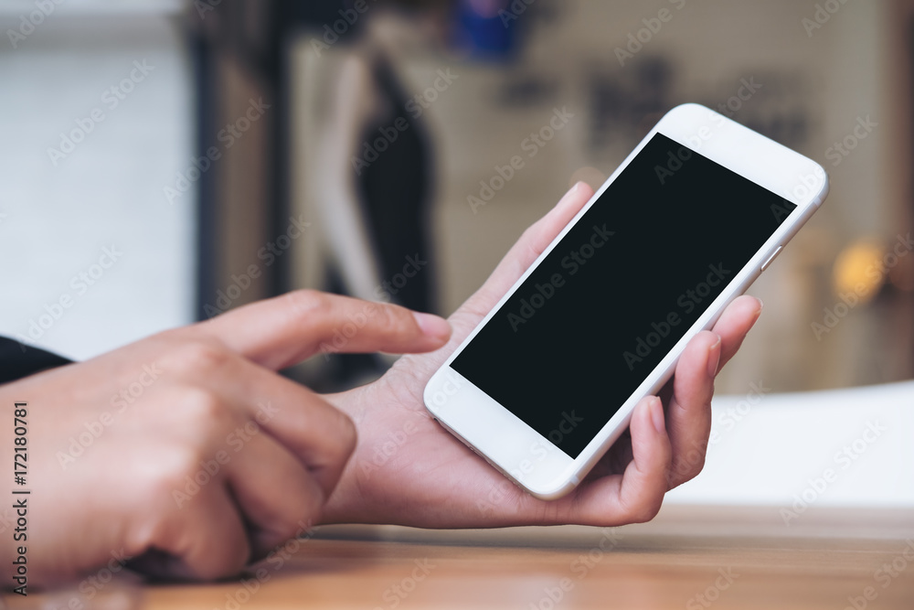 Mockup image of a woman's hand holding and pointing at white mobile phone with blank black screen in modern cafe