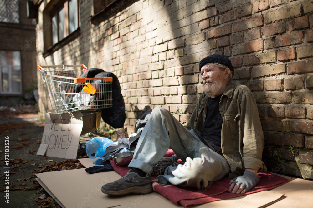 Homeless man sitting on cardboard by the brick wall. Tramp on the ground,  shopping cart with his belongings standing close. Photos | Adobe Stock