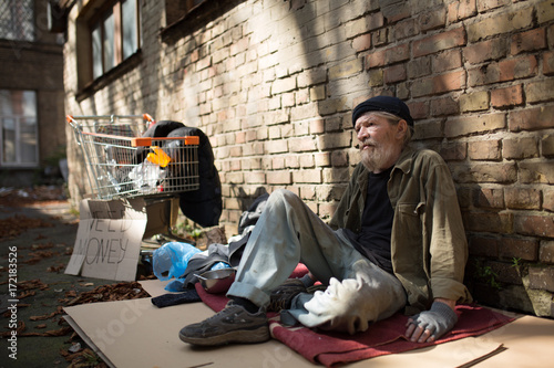 Homeless man sitting on cardboard by the brick wall. Tramp on the ground, shopping cart with his belongings standing close.