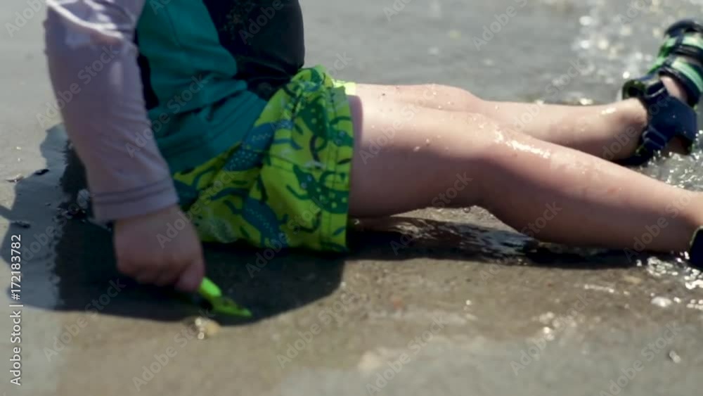 Little Boy Digs In Sand With Shovel, Gentle Waves Lap Over His Legs, Slow Motion Closeup Little Boy Digs In Sand With Shovel, Gentle Waves Lap Over His Legs, Slow Motion Closeup