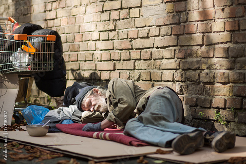 Sleeping homeless man lying on cardboard. Tramp lying by the brick wall, his posessions on the ground and in shopping cart.