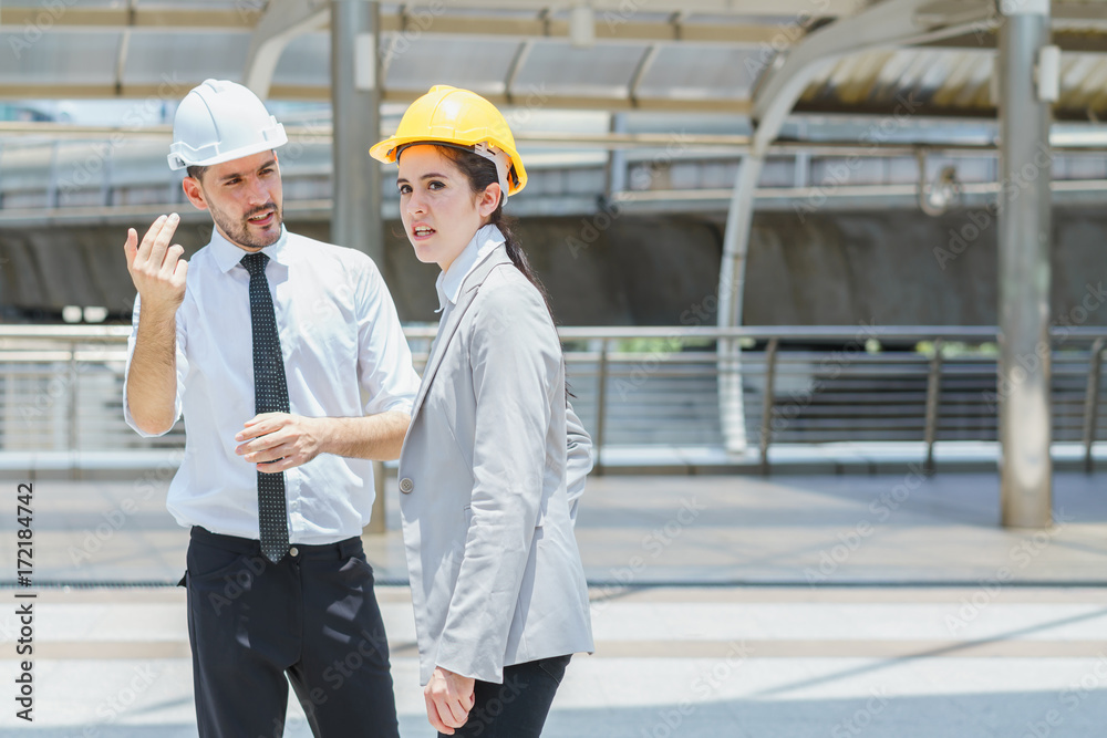 Two engineer look to target at site outdoors. Couple wearing safety helmet with city background. Concept of business partnership meeting.