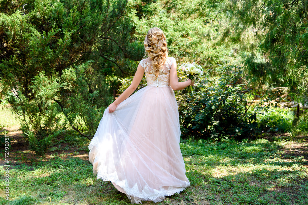 Beautiful bride in wedding dress with bridal bouquet dancing in the park outdoors, back view. Blond girl with curly hair styling and jewelry, free space. Full length body portrait