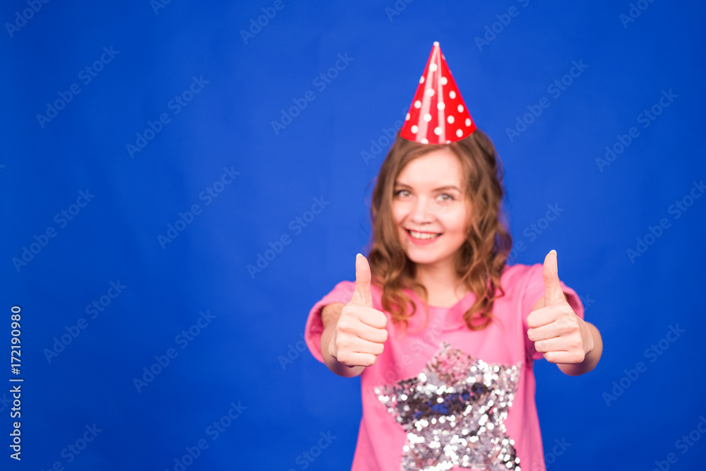 Celebration and party. Having fun. Young pretty woman in birthday hat is laughing and showing thumbs up. Colorful studio portrait with blue background. Celebration and party. Having fun. Young pretty woman in birthday hat is laughing and showing thumbs up. Colorful studio portrait with blue background.