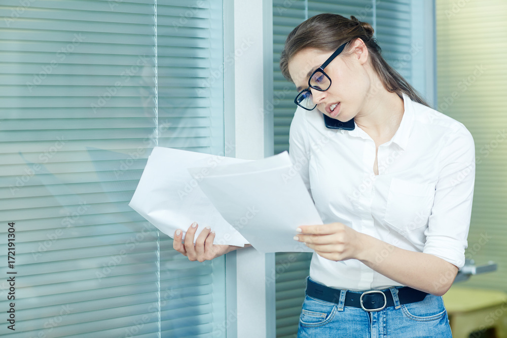 Busy young manager in eyeglasses analyzing financial figures while talking to her colleague on smartphone, waist-up portrait Busy young manager in eyeglasses analyzing financial figures while talking to her colleague on smartphone, waist-up portrait