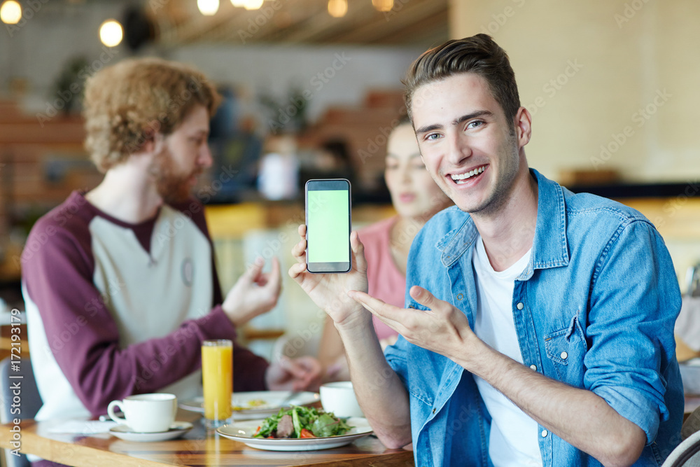 Handsome young man with wide smile posing for photography while pointing at blank screen of modern smartphone, his friends chatting animatedly with each other and enjoying lunch at cozy cafe Handsome young man with wide smile posing for photography while pointing at blank screen of modern smartphone, his friends chatting animatedly with each other and enjoying lunch at cozy cafe