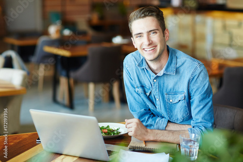 Waist-up portrait of handsome young journalist looking at camera with wide smile while working on article and enjoying delicious lunch at cozy small cafe