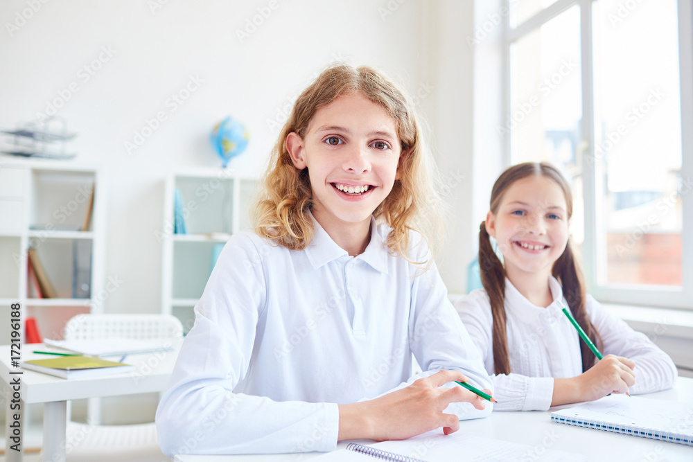 Friendly classmates with pencils looking at camera in classroom Friendly classmates with pencils looking at camera in classroom