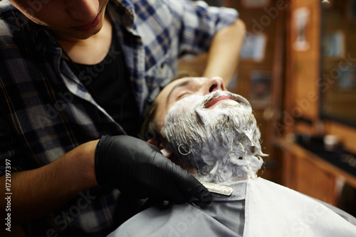 Fotografie Bearded man being shaven with sharp dangerous razor by professional barber
