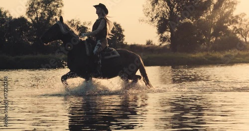 Slow motion 240fps shot of cowboy girl riding horse in water of sunset river