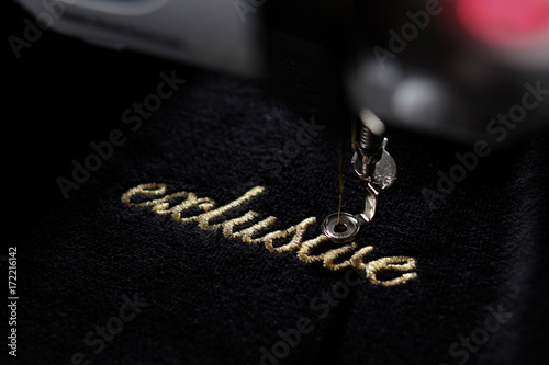 embroidery of gold lettering "exclusive" on black velvetely fabric with embroidery machine - diagonal view with part of machine - background and foreground blanked out blurry 