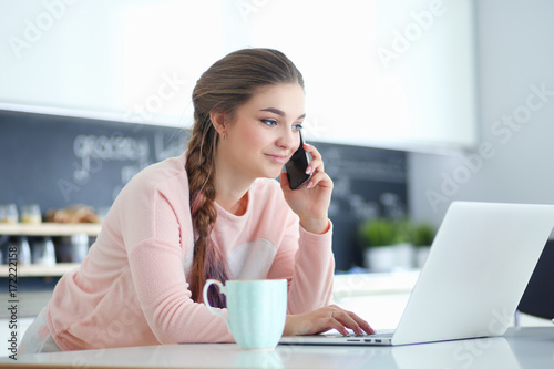 Young woman sits at the kitchen table using a laptop and talking on a cell phone.