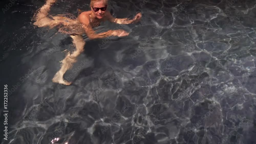Beautiful, slim, caucasian woman wearing string bikini getting into private pool lined with flowers and grey wall. Camera moves with swimmer.