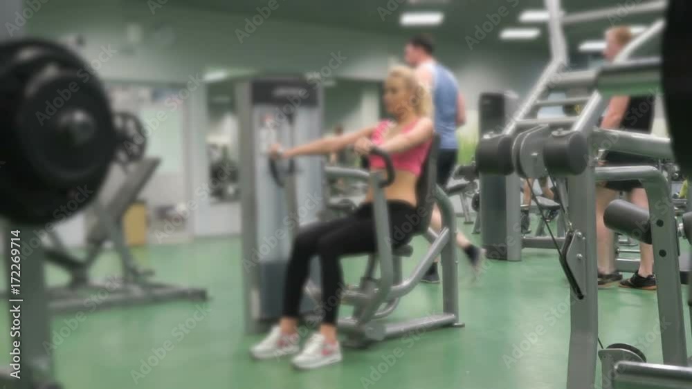 Sports workout in the gym indoors. Young beautiful blond girl in sport suit training her arms on the sports simulator