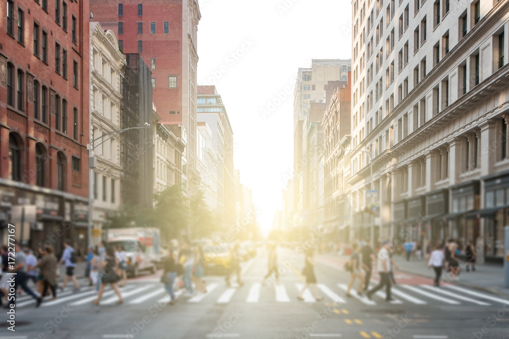 Groups of people walking across a busy crosswalk intersection in New York City with the glow of the sun in the background