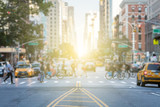 People crossing the busy intersection between traffic on 3rd Avenue and 10th Street in Manhattan in New York City with the glow of sun light in the background