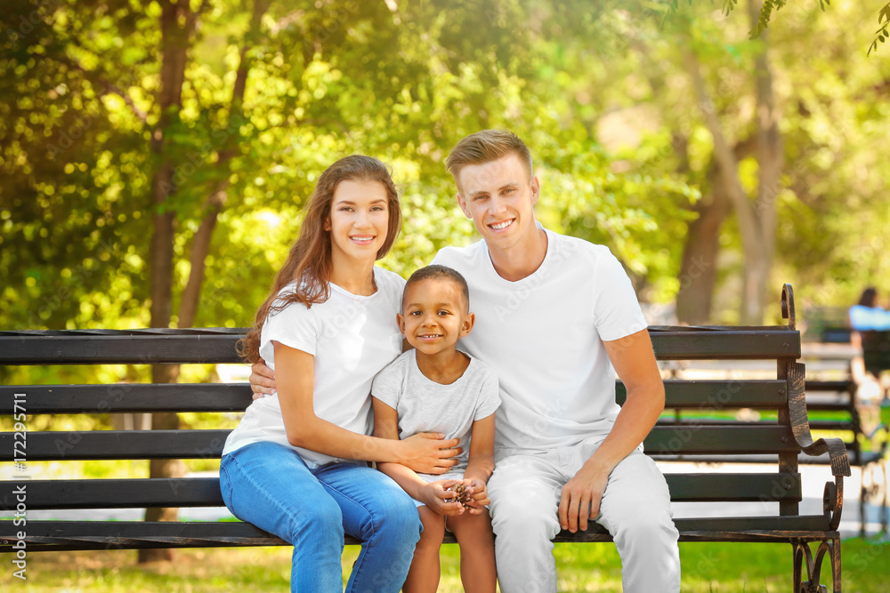Young family with adopted African American boy sitting on bench in park Young family with adopted African American boy sitting on bench in park