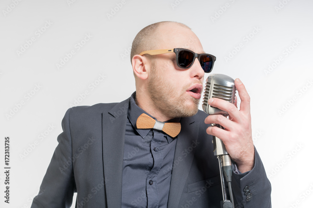 Naklejka premium Unshaven bald man in a gray suit and sunglasses holding a microphone and singing. Toned