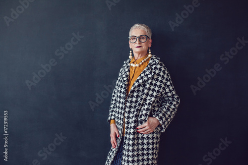 Stylish old woman in glasses standing on a black background.