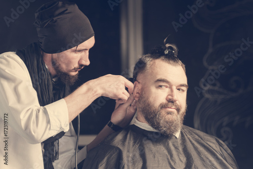 Hairdresser in a black cap and a black apron cuts with hairclipper a man with a clip on the head