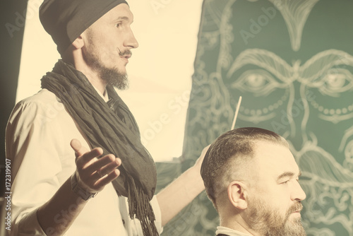 Man hairdresser in a white shirt, black hat and scarf doing hairstyle a bearded man