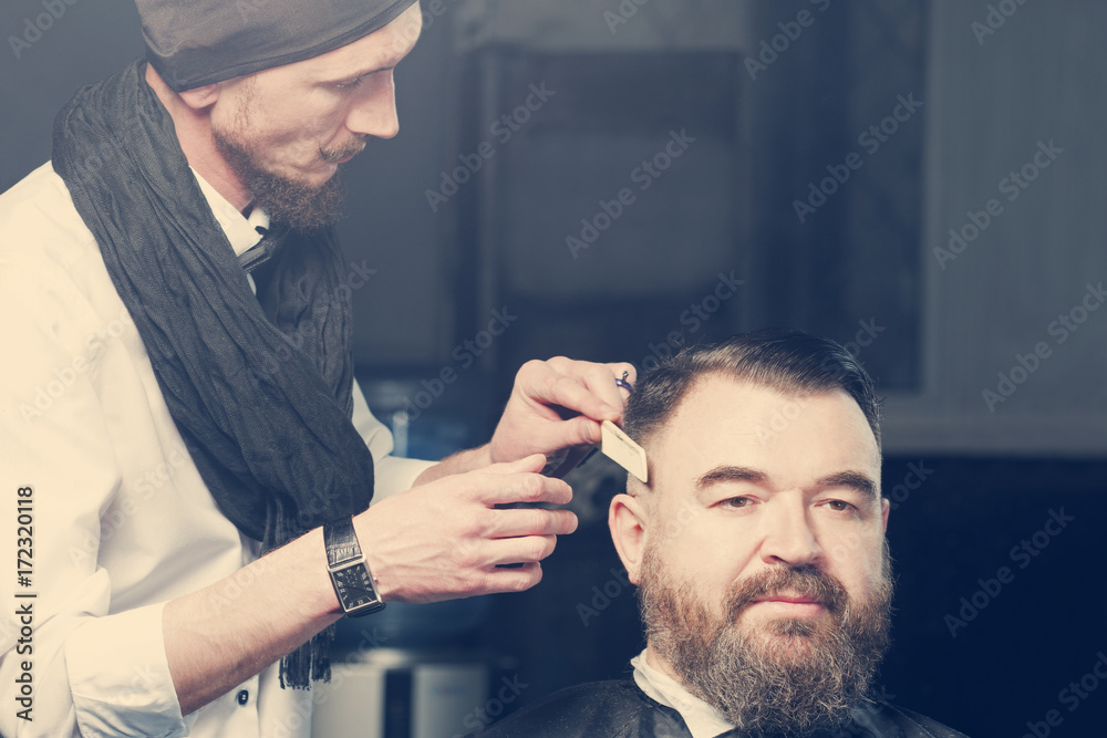 Stylist in black hat, bow tie and scarf makes hair a man with a beard Stylist in black hat, bow tie and scarf makes hair a man with a beard