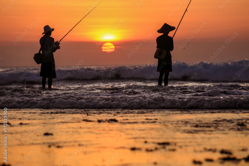 Silhouette of fishermen on the quiet ocean with the rays of sunset at Jimbaran beach, Bali, Indonesia 