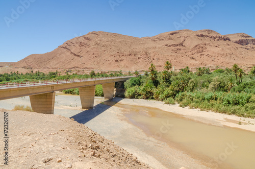 Bridge spanning over dry river bed with some water, mountains and palms in Morocco, North Africa