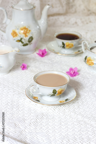 A tea cup decorated with flowers, two biscuits and a candle on a white wooden table. In the background a turquoise clock on a yellow wainscot. Vintage tea time