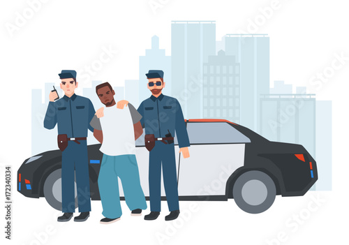 Two policemen in uniform standing near police car with caught criminal against city buildings on background